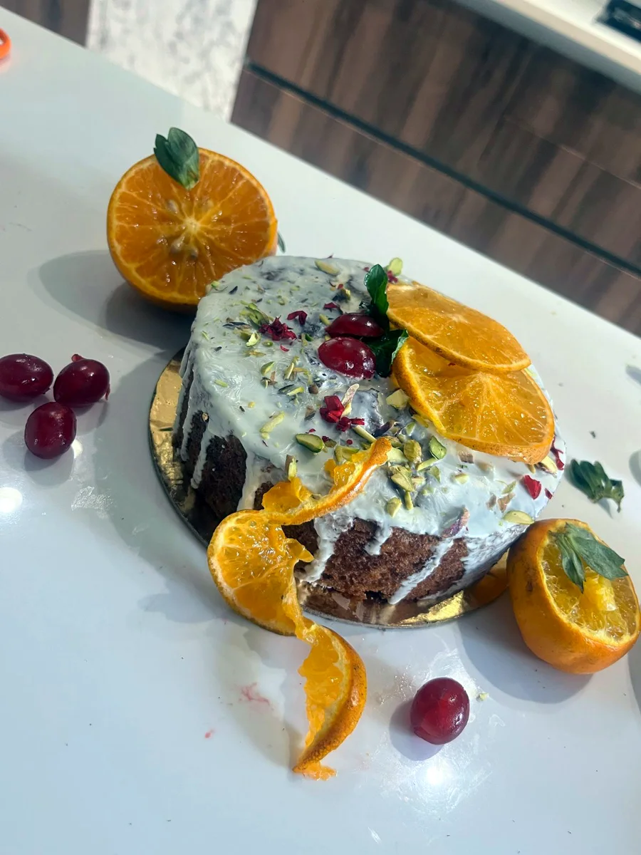 An orange tea cake with white chocolate glaze, pistachios, rose petals, cherries and fresh orange slices