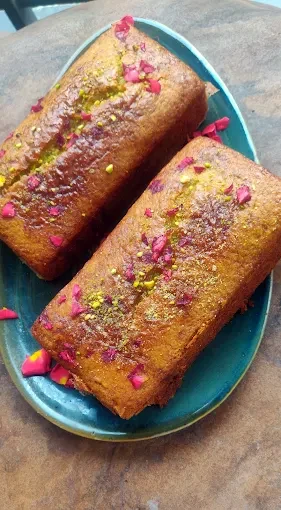 Two rose and pistachio loaf cakes topped with rose petals and pistachios on a teal platter