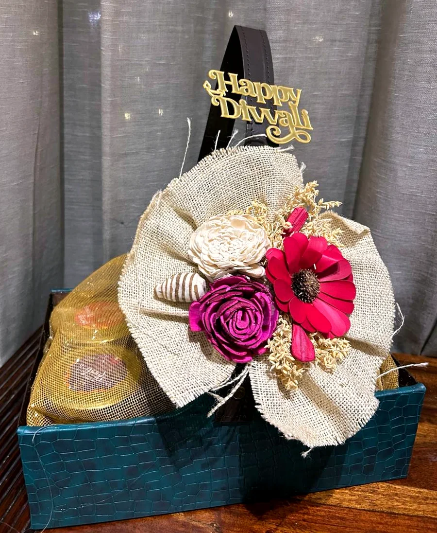 A teal leather-finish hamper basket with a jute bow, sola wood flowers and a Happy Diwali topper